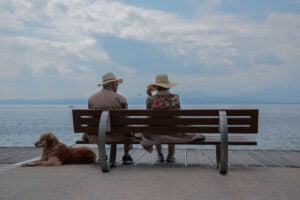 Two elderly people sitting on a bench near the sea with a dog laying nearby